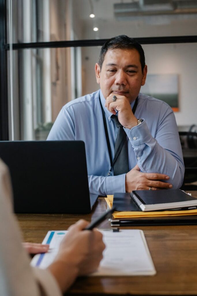 pexels-photo-5668852 Serious mature ethnic entrepreneur sitting at wooden table with documents and laptop and thinking while crop anonymous colleague taking notes on clipboard